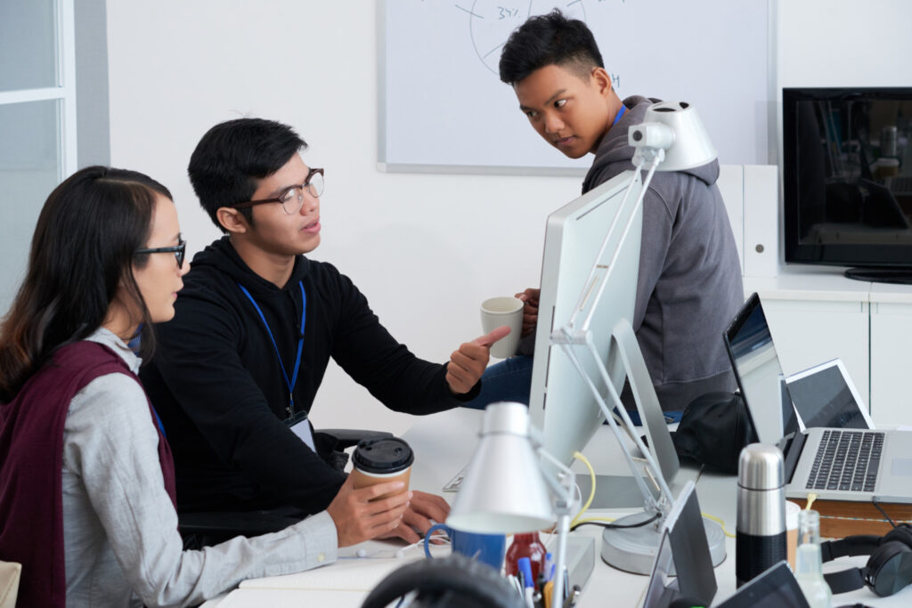Three young adults engaged in a discussion around a computer in a modern office, with one person pointing at the monitor while holding a coffee cup.