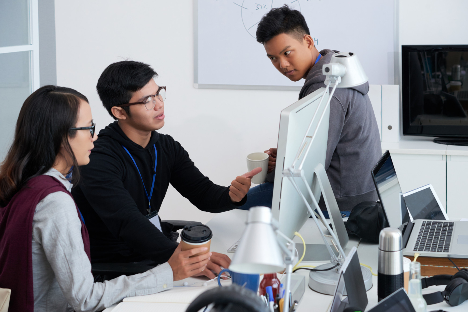 Three young adults engaged in a discussion around a computer in a modern office, with one person pointing at the monitor while holding a coffee cup.
